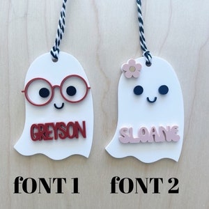 May include: Two white ghost ornaments with black and white string. The ghost on the left has red glasses and the name "Greyson" in red letters. The ghost on the right has a pink flower and the name "Sloane" in pink letters. The text "FONT 1" is below the left ghost and "FONT 2" is below the right ghost.