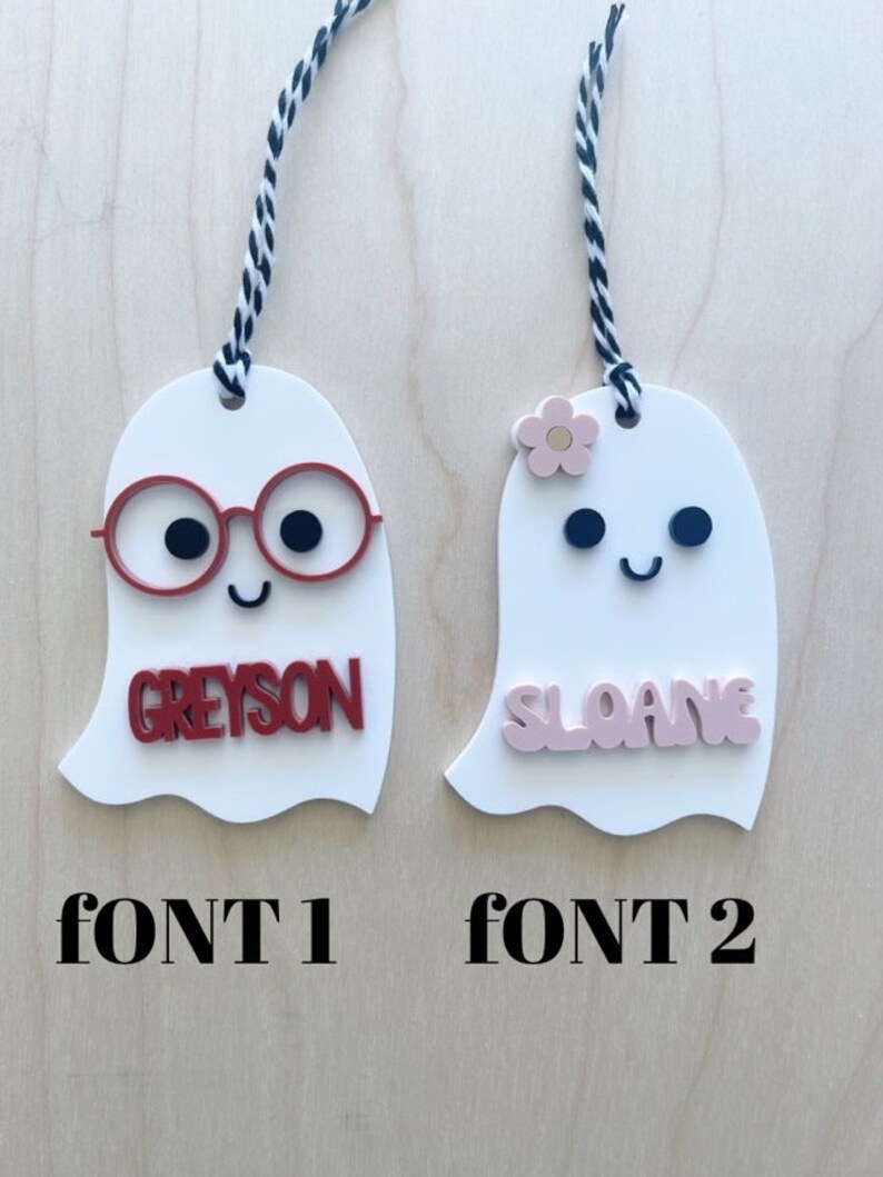 May include: Two white ghost ornaments with black and white string. The ghost on the left has red glasses and the name "Greyson" in red letters. The ghost on the right has a pink flower and the name "Sloane" in pink letters. The text "FONT 1" is below the left ghost and "FONT 2" is below the right ghost.