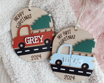 Puede incluir: Dos adornos navideños de madera con forma de camiones que transportan árboles de Navidad. El camión de la izquierda es rojo y dice "Feliz Navidad" y "Grey" con el año "2024". El camión de la derecha es azul y dice "Mi primera Navidad" y "Lukas" con el año "2024".