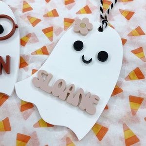 May include: White ghost-shaped ornament with a pink flower, black eyes, and a black smile. The ornament has the name "Sloane" written in pink letters.