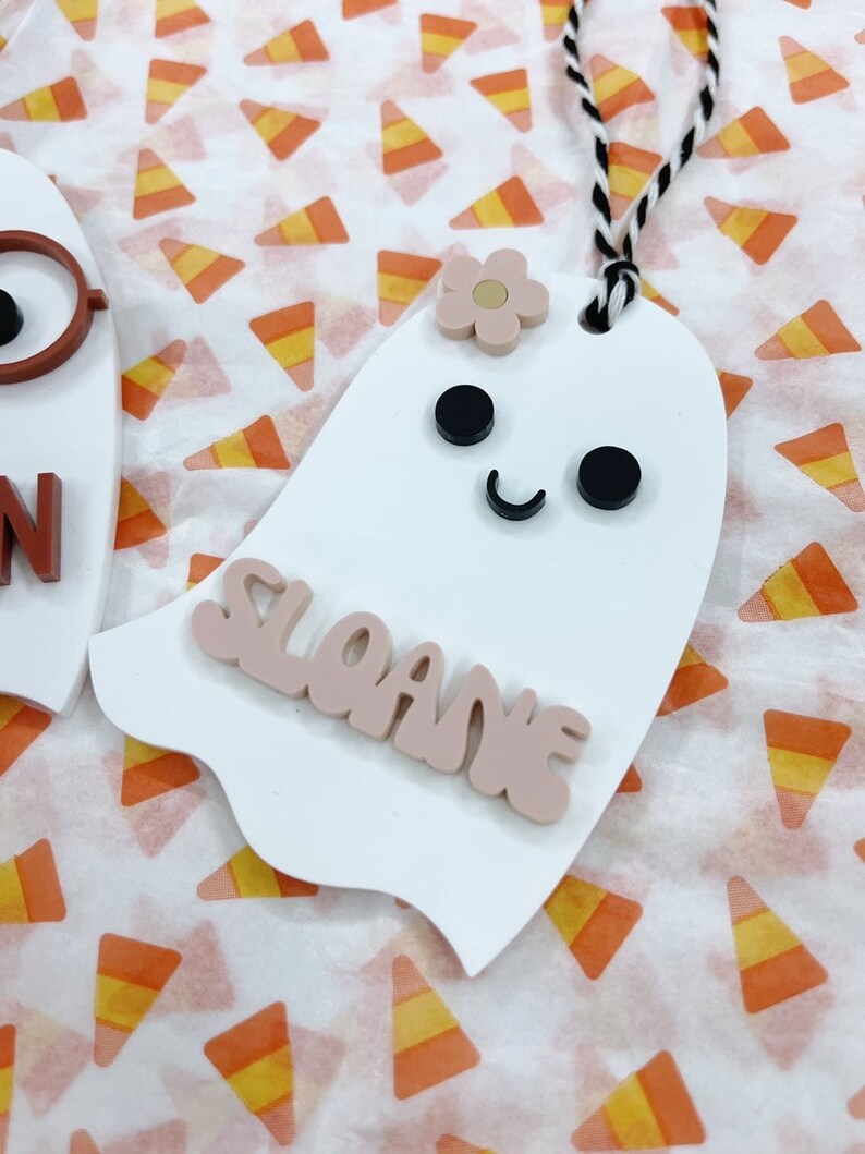 May include: White ghost-shaped ornament with a pink flower, black eyes, and a black smile. The ornament has the name "Sloane" written in pink letters.