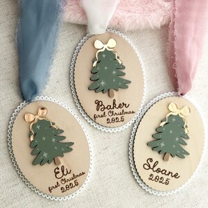May include: Three wooden Christmas ornaments with a green tree design and gold bow accents. Each ornament has a name and the text "first Christmas 2025". The ornaments are oval-shaped with a scalloped edge and are hanging from ribbons.