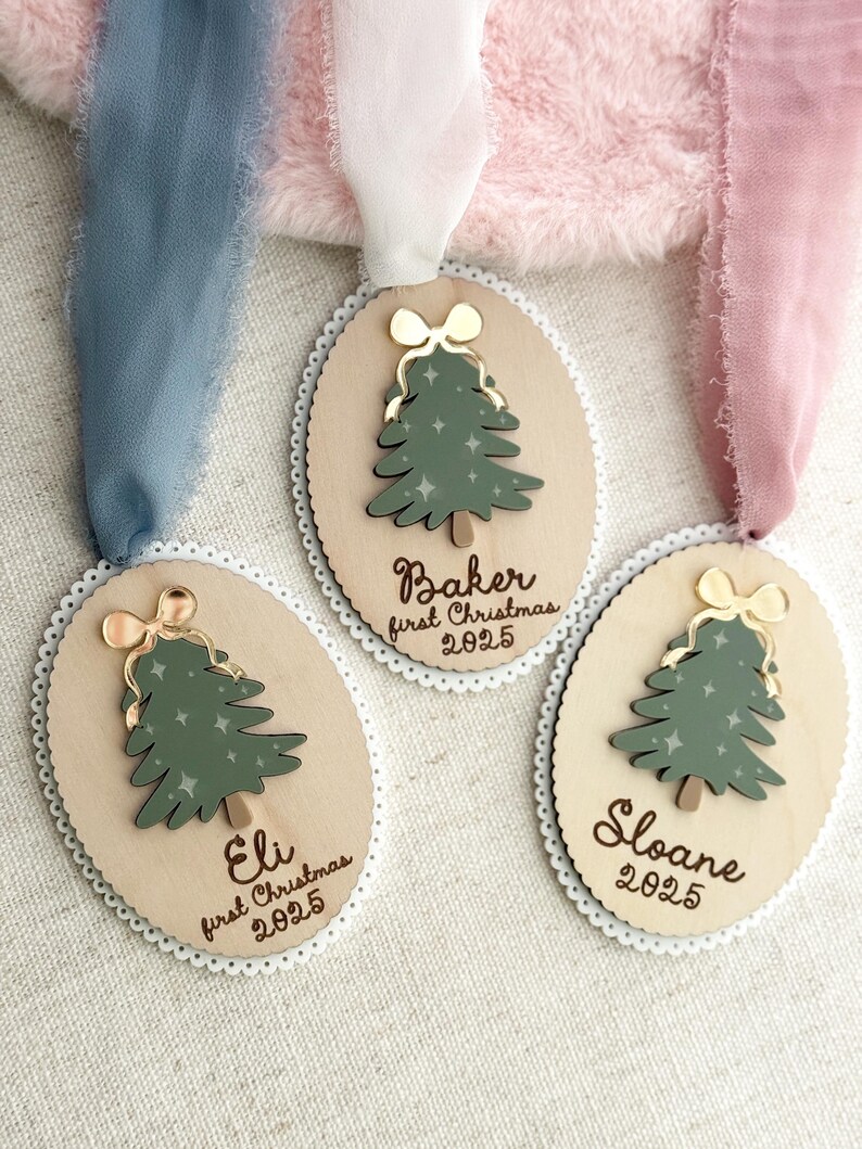 May include: Three wooden Christmas ornaments with a green tree design and gold bow accents. Each ornament has a name and the text "first Christmas 2025". The ornaments are oval-shaped with a scalloped edge and are hanging from ribbons.