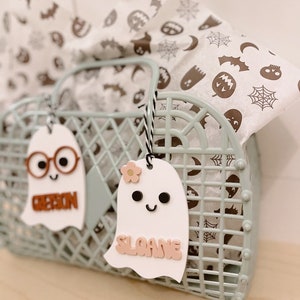 May include: Two white ghost-shaped gift tags with names "Ribbon" and "Sloane" written in pink letters. The tags are hanging from a light green plastic basket filled with white tissue paper with a Halloween pattern.
