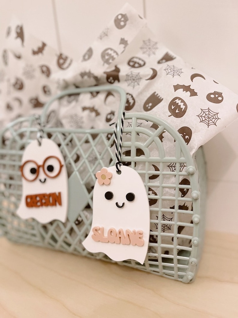 May include: Two white ghost-shaped gift tags with names "Ribbon" and "Sloane" written in pink letters. The tags are hanging from a light green plastic basket filled with white tissue paper with a Halloween pattern.