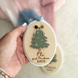 May include: Wooden Christmas ornament with a green tree design, gold bow, and the text "Eli first Christmas 2025." The ornament has a white lace trim and a blue ribbon for hanging.