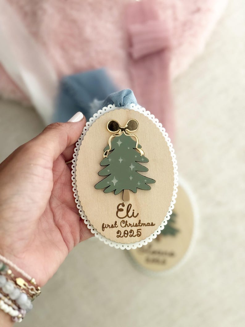 May include: Wooden Christmas ornament with a green tree design, gold bow, and the text "Eli first Christmas 2025." The ornament has a white lace trim and a blue ribbon for hanging.