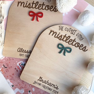 Puede incluir: Dos letreros de madera con el texto "The Cutest Little Mistletoes" en letra cursiva marr&oacute;n. El letrero de arriba tiene un lazo rojo y el texto "Alessa First Christmas". El letrero de abajo tiene un lazo verde y el texto "Graham's Second Christmas 2024".
