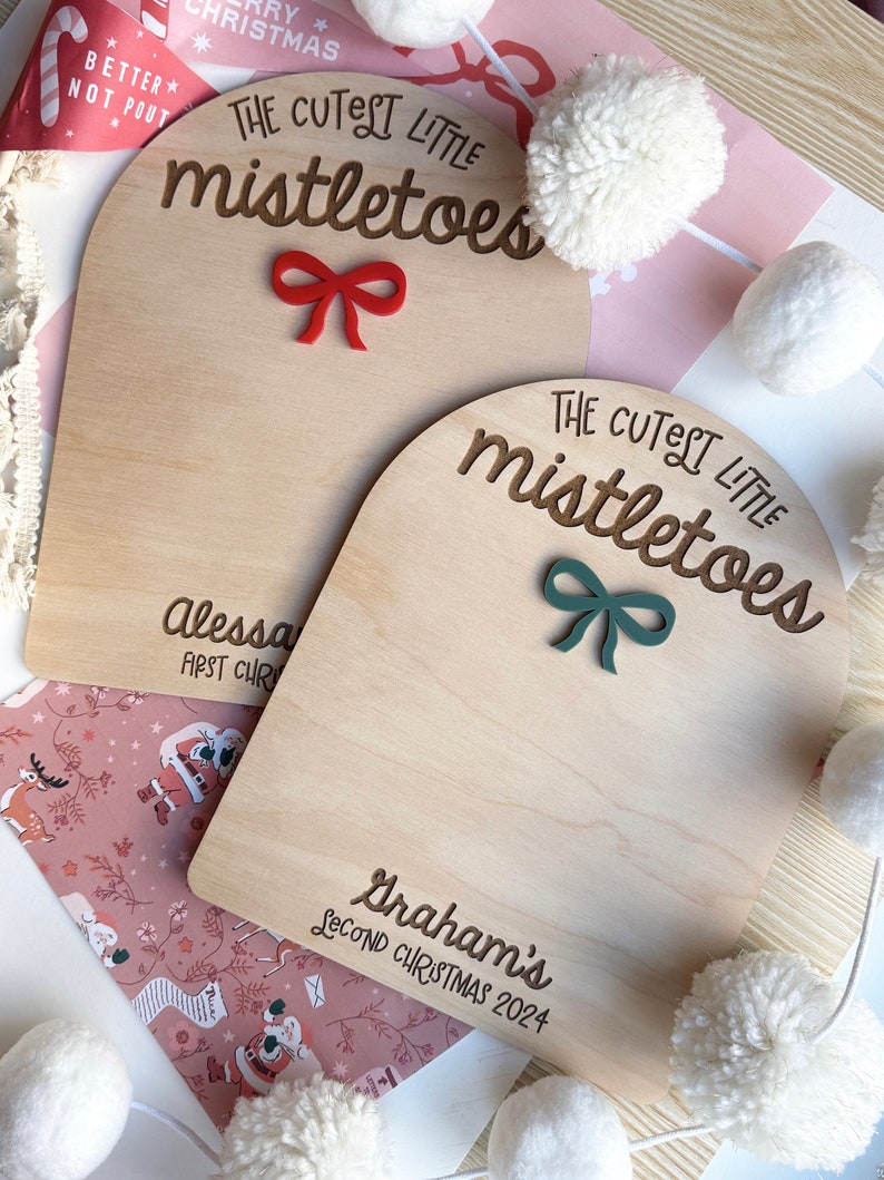 Puede incluir: Dos letreros de madera con el texto "The Cutest Little Mistletoes" en letra cursiva marr&oacute;n. El letrero de arriba tiene un lazo rojo y el texto "Alessa First Christmas". El letrero de abajo tiene un lazo verde y el texto "Graham's Second Christmas 2024".