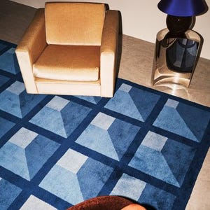 Könnte beinhalten: Ein beiger Polstersessel steht auf einem blauen Teppich mit geometrischem Gittermuster. Neben dem Sessel befindet sich ein metallischer Beistelltisch mit dunklem Fuß und einem leuchtend blauen Lampenschirm. Ein Teil eines dunkelorangen Möbelstücks ist sichtbar.