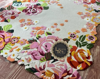 May include: A floral rug with a cream backdrop and a border of colourful flowers. The rug features large, detailed roses in shades of pink, orange, and yellow, with green leaves. The rug is placed on a wooden floor.