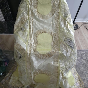 May include: Cream-colored garment with yellow circular accents and intricate gold embroidery. The fabric features a delicate lace trim along the edges. The design includes a series of radiating lines and circular patterns, creating a textured appearance.