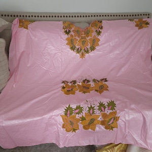 May include: A light pink garment with intricate floral embroidery in shades of gold, green, and pink. The embroidery is concentrated around the neckline and lower portion of the garment, creating a decorative pattern. The fabric has a subtle textured appearance.