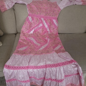 May include: A pink dress with long, flared sleeves and intricate embroidery. The dress features a high neckline and a flowing skirt with multiple tiers. The fabric has a shiny appearance, and the dress is adorned with delicate lace trim.