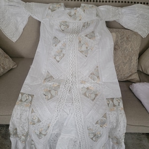 May include: A white, long-sleeved dress with intricate lace and embroidered details. The dress features a flowing silhouette with bell sleeves and decorative panels. The design incorporates paisley patterns and delicate trim, creating a bohemian aesthetic.