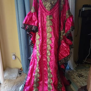 May include: A long, pink dress with gold embroidery and black lace trim. The dress has a V-neck and long sleeves.