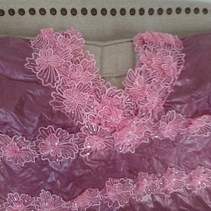 May include: A mauve fabric with a shiny, textured appearance. Pink floral lace trim with sequins and bead accents adorns the neckline and edges of the fabric. The lace features detailed flower designs.