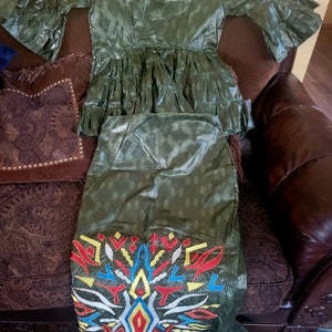 May include: A two-piece outfit in a green fabric with a geometric pattern. The top has a ruffled hem and short sleeves. The skirt has a colorful embroidered design.