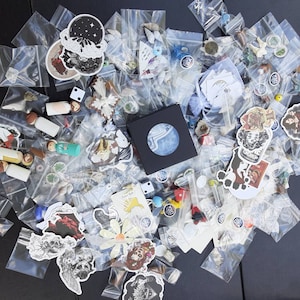 May include: A collection of small items, including stickers, dice, wooden dolls, and various trinkets, are contained in clear plastic bags. A black box with a circular design is also present. The items are scattered on a dark surface.