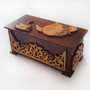 Puede incluir: Una caja de madera con una tapa decorativa que presenta una tetera y una taza de té talladas. La caja tiene un acabado marrón y recortes intrincados alrededor de los bordes.