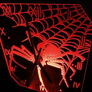 Può includere: Un orologio da parete rosso e illuminato con un design a ragno e ragnatela. Il quadrante dell'orologio presenta numeri romani e lancette. Il ragno e la ragnatela sono finemente dettagliati, creando un'estetica spettrale a tema Halloween.