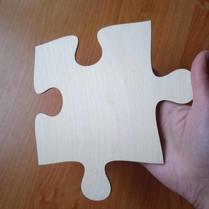 May include: A single, unfinished wooden jigsaw puzzle piece. The piece is held in a hand and is light-coloured with a natural wood grain. The background is a wooden surface.