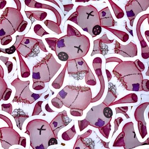 Può includere: Un mucchio di adesivi rosa e bianchi con un'illustrazione di un cartone animato di una bambola cucita, rotta, con una croce sulla fronte. La bambola ha un naso a bottone, un cuore sul petto e una coda cucita.