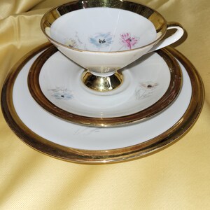 Könnte beinhalten: Ein weißes Tee-Set mit goldfarbenem Rand und Blumenmuster. Die Tasse hat eine breite, flache Schale und einen Henkel. Die Untertasse hat einen erhöhten Rand und ein Blumenmuster.