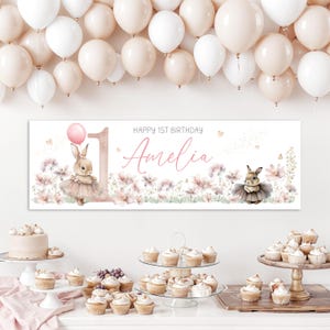 May include: A banner for a first birthday party with the name "Amelia" in cursive script. The banner features a bunny wearing a tutu, a pink balloon, and floral accents. White and peach balloons are in the background.