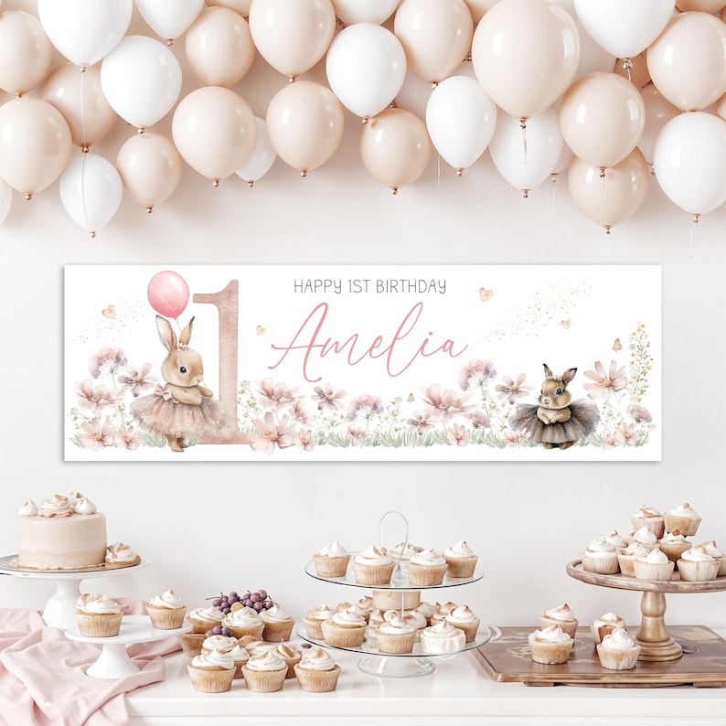 May include: A banner for a first birthday party with the name "Amelia" in cursive script. The banner features a bunny wearing a tutu, a pink balloon, and floral accents. White and peach balloons are in the background.