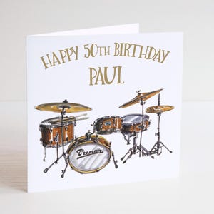 May include: A white birthday card featuring a watercolour illustration of a drum kit. The card has gold text reading "HAPPY 50TH BIRTHDAY PAUL". The drum set includes brown drums, cymbals, and the Premier logo.