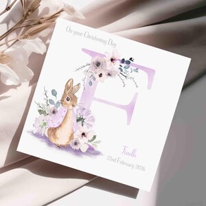 Peut inclure: Carte de baptême carrée avec l'inscription "On your Christening Day". Elle présente une illustration à l'aquarelle d'un lapin, de fleurs et de la lettre "F". Les couleurs sont douces, avec du rose et du violet.