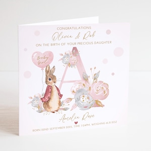 May include: A pink baby announcement card featuring a bunny illustration holding a heart-shaped balloon. The card reads "Congratulations Olivia & Rob on the birth of your precious daughter." The card also includes the baby's name and birth details.