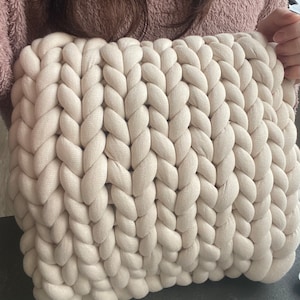 Può includere: Un cuscino grande di colore crema lavorato a maglia con una trama intrecciata. Il cuscino è realizzato in filato spesso e morbido e ha un aspetto spesso e accogliente.