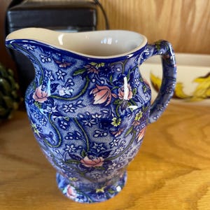 Peut inclure: Une cruche en céramique bleue et blanche avec un motif floral. La cruche présente un bord et une anse de couleur crème. Des fleurs roses et jaunes sont parsemées dans le motif. La cruche mesure environ 18 cm de haut.