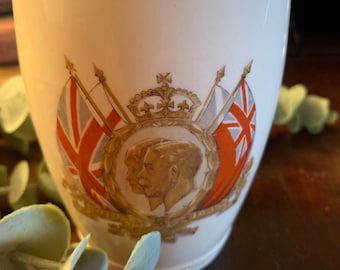 Puede incluir: Una taza de cerámica blanca con una banda dorada y las palabras "Silver Jubilee" impresas en ella. La taza presenta una imagen colorida de una corona, dos banderas y un retrato de perfil de un rey y una reina.