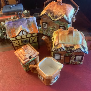 May include: A set of four ceramic teapots shaped like cottages with thatched roofs. The teapots are decorated with brown, green, yellow, and white paint. The teapots are arranged on a red surface.