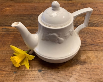 May include: A white ceramic teapot with a lid and handle, featuring embossed fruit and leaf designs. A yellow daffodil rests beside the teapot on a wooden surface. The teapot has a spout and a rounded base.
