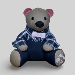 May include: A grey teddy bear wearing a blue plaid shirt, navy blue overalls, and a light pink bow tie. The bear has pink inner ears, a black nose, and a decorative patch on its leg.