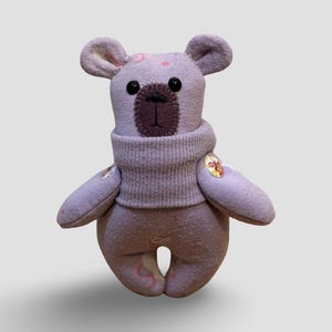 May include: A handmade lavender teddy bear with black button eyes and a brown stitched nose. The bear wears a matching lavender sweater and has small, round ears. A small, colourful patch is sewn on the bear's arm.
