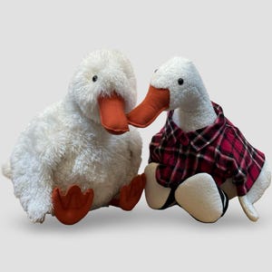 May include: Two plush white duck toys with orange beaks and feet. One duck is wearing a red and black plaid shirt. The ducks are sitting side by side against a light grey background. These stuffed animals are perfect for a child's bedroom or nursery.