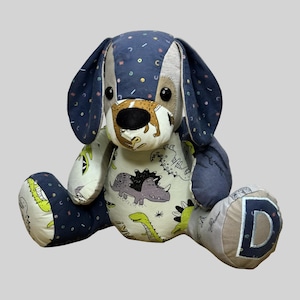 May include: A handmade stuffed dog toy with blue and tan patterned fabric. The dog features large floppy ears, a tan snout, and a body covered in dinosaur and polka dot patterns. The letter "D" is sewn on one paw.