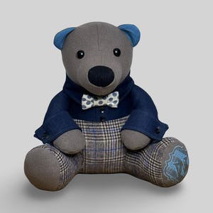 May include: A grey teddy bear wearing a blue denim jacket, a plaid waistcoat, and a white bow tie with black polka dots. The bear has blue ear accents and black button eyes. The bear's feet have a blue embroidered design.