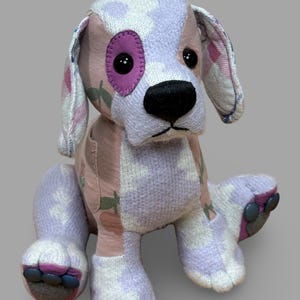 May include: A handmade plush dog with a patchwork design. The dog features a mix of pink, lavender, and white fabrics with a floral pattern. It has a purple eye patch, black button eyes, and a black nose. The dog is sitting with its paws forward.