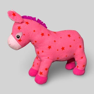 May include: A pink donkey soft toy decorated with red stars. The toy has a pink mane, ears, and hooves. The soft toy is made from fabric and is designed for children.
