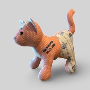 May include: A peach and white stuffed cat toy. The cat is made from peach fabric with white polka dots, and the sides have a striped pattern with seashell motifs. The text "Маргарита 05.09.1989" is embroidered on the side of the toy.