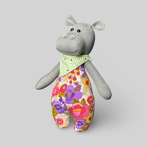 May include: A grey stuffed hippo toy wearing floral patterned overalls and a green polka-dot bandana. The overalls have a vibrant pattern of purple, pink, and yellow flowers. The hippo has button eyes and grey arms and legs.