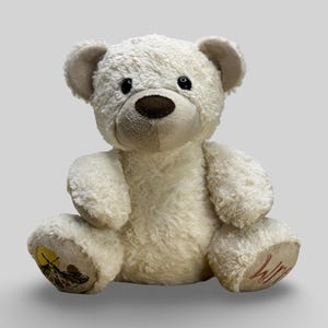 May include: A cream teddy bear with a brown nose and black eyes. The bear has a soft, plush texture and is sitting with its legs outstretched. The bear's paw pads have embroidered details, including the word "Win".