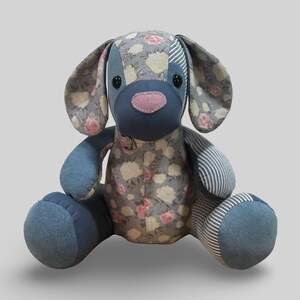 May include: A handmade patchwork dog toy. The dog has a gray floral patterned body, blue denim accents, and striped fabric on the ears and paws. It has a pink nose and black button eyes. A charming, soft toy.