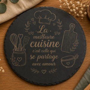 Peut inclure: Un plateau de service rond en ardoise noire gravé d'un motif représentant une toque de chef, un pot avec des ustensiles, une planche à découper et un bol. Le texte indique : La meilleure cuisine c'est celle qui se partage avec amour.